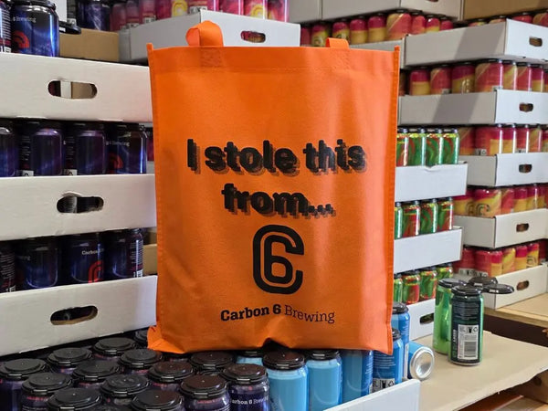 Stolen tote bag in orange with the text 'I stole this from...' displayed, set among beer cans from Carbon 6 Brewing.
