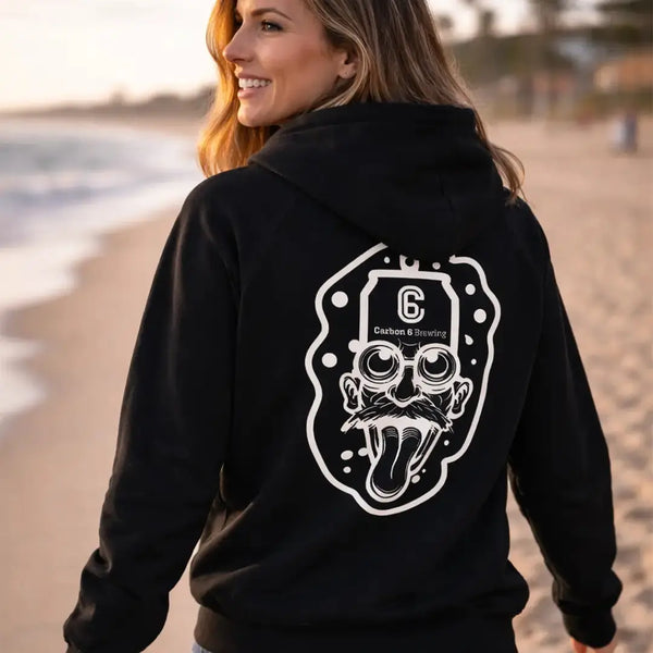Woman wearing a Carbon 6 Can Head Brewer Fleece Hoodie with a graphic design on the back, standing on the beach.