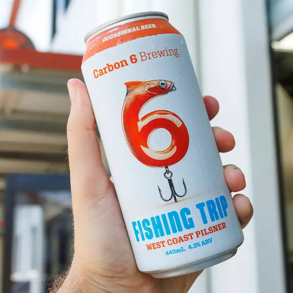 A person holding a can of The Occasional Beer with a design featuring fishing themes and the label 'Fishing Trip'.