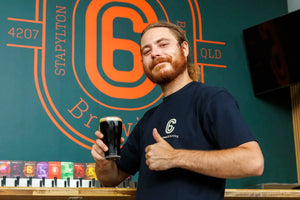 new brewer at Carbon 6 Brewing proudly holding a pint of dark beer in front of the brewery's logo.